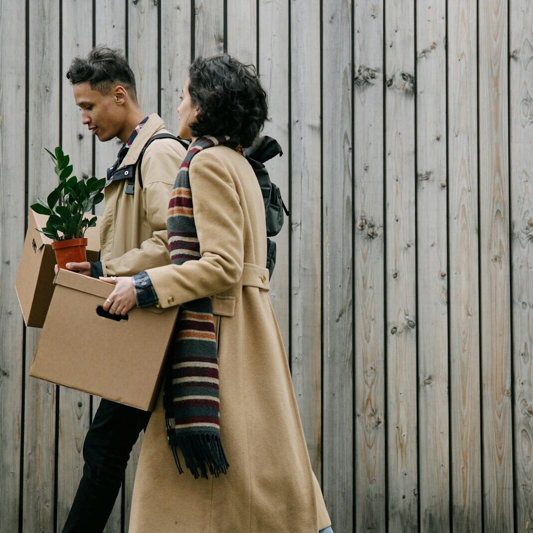 Couple walking with boxes and a potted plant, wearing winter coats, outdoors.